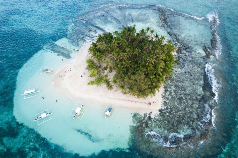 Aerial view of a tiny palm-covered island ringed by reef and bangka boats