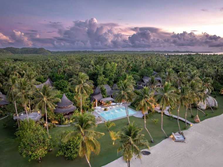 Aerial view of a resort pool and thatched pavilions under a violet sunset sky