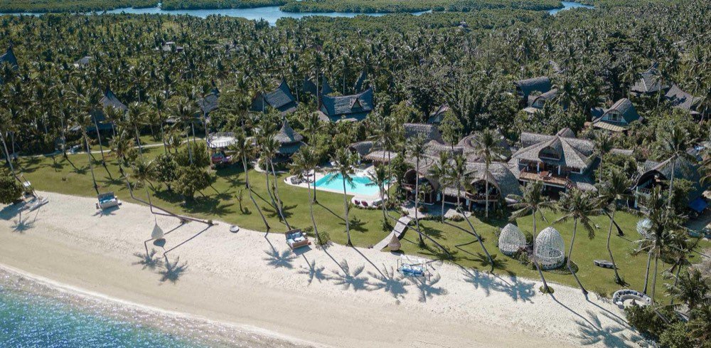 Aerial view of a thatched-roof luxury resort set among coconut palms along a white-sand Siargao beach